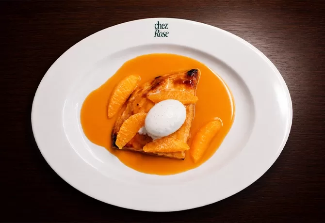 A slice of orange tart topped with a scoop of cream sits on a white plate, surrounded by orange sauce and orange segments, with chez Rose written at the top of the plate.