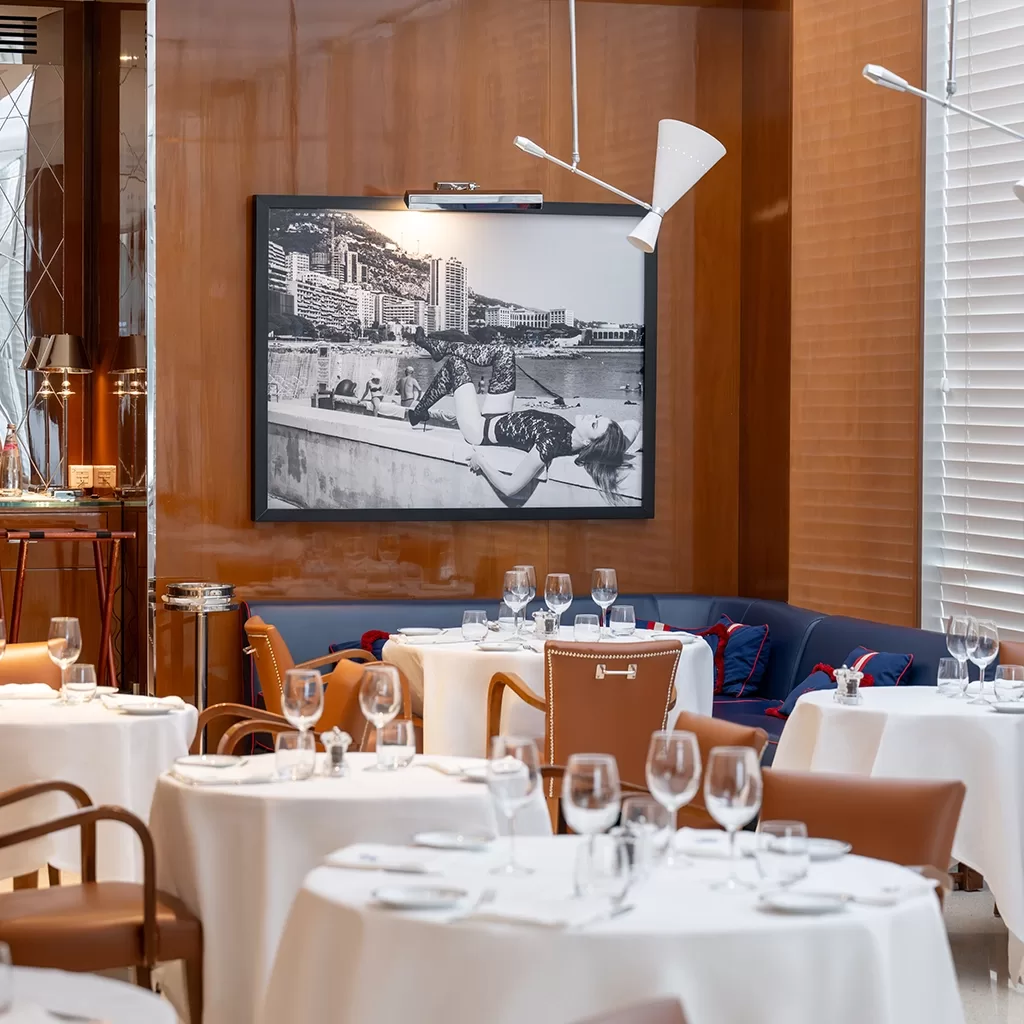 Elegant restaurant dining area with round tables set with glassware and white tablecloths, brown and blue seating, wood-paneled walls, modern white light fixtures, and a large black-and-white photo on the wall.