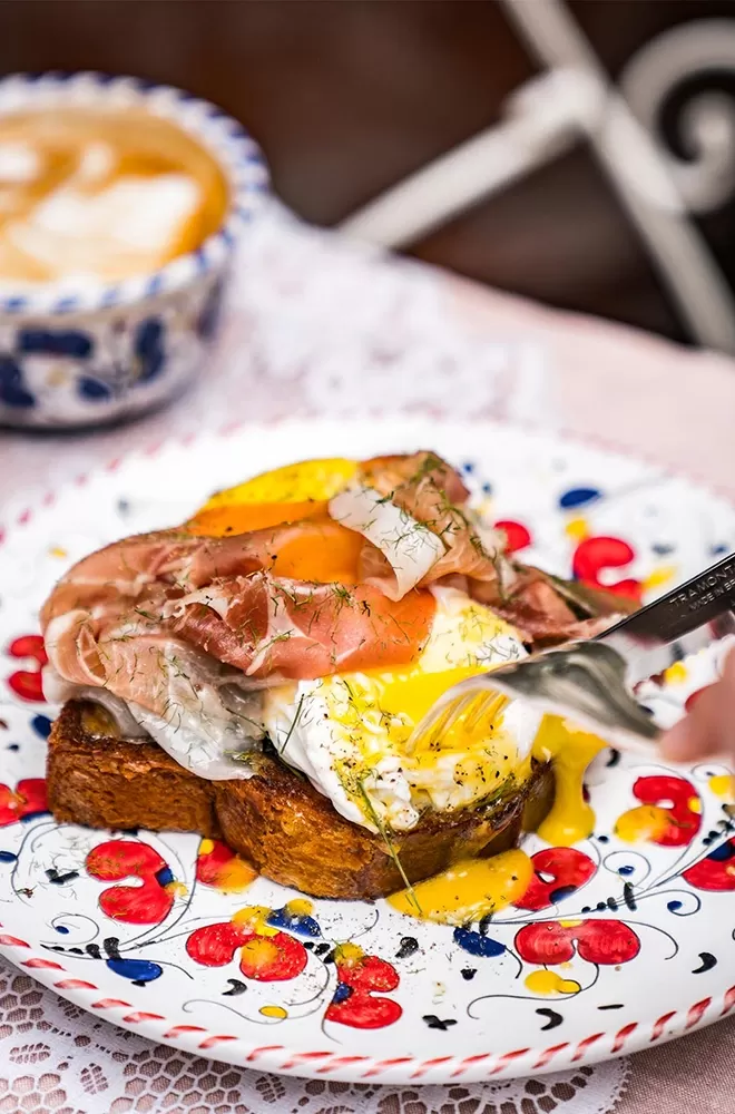 A vibrant plate with toasted bread topped with poached eggs, ham, and seasonings. A fork cuts into the egg, letting the yolk drip down. In the background, theres a coffee cup with a decorative pattern at Circolo Popolare, London.
