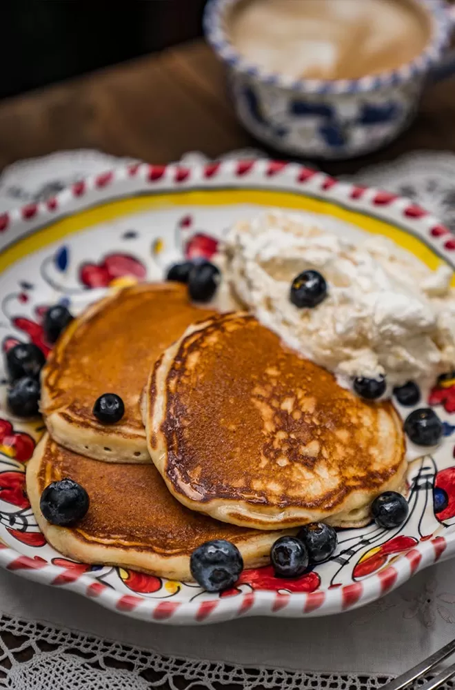 A decorative plate with three stacked pancakes topped with blueberries. A dollop of cream sits beside the pancakes. The plate features a floral pattern and is set on a wooden table with a lace tablecloth. A blurred cup is in the background at Circolo Popolare, London.