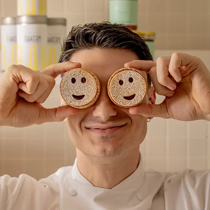 A smiling person holds two round cookies with smiling faces in front of their eyes, standing in a kitchen.