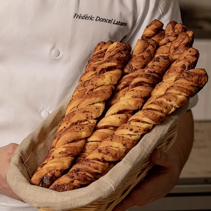 A person in a white chef’s coat holds a wicker basket lined with cloth, filled with several long, twisted breadsticks with a golden, crispy texture.
