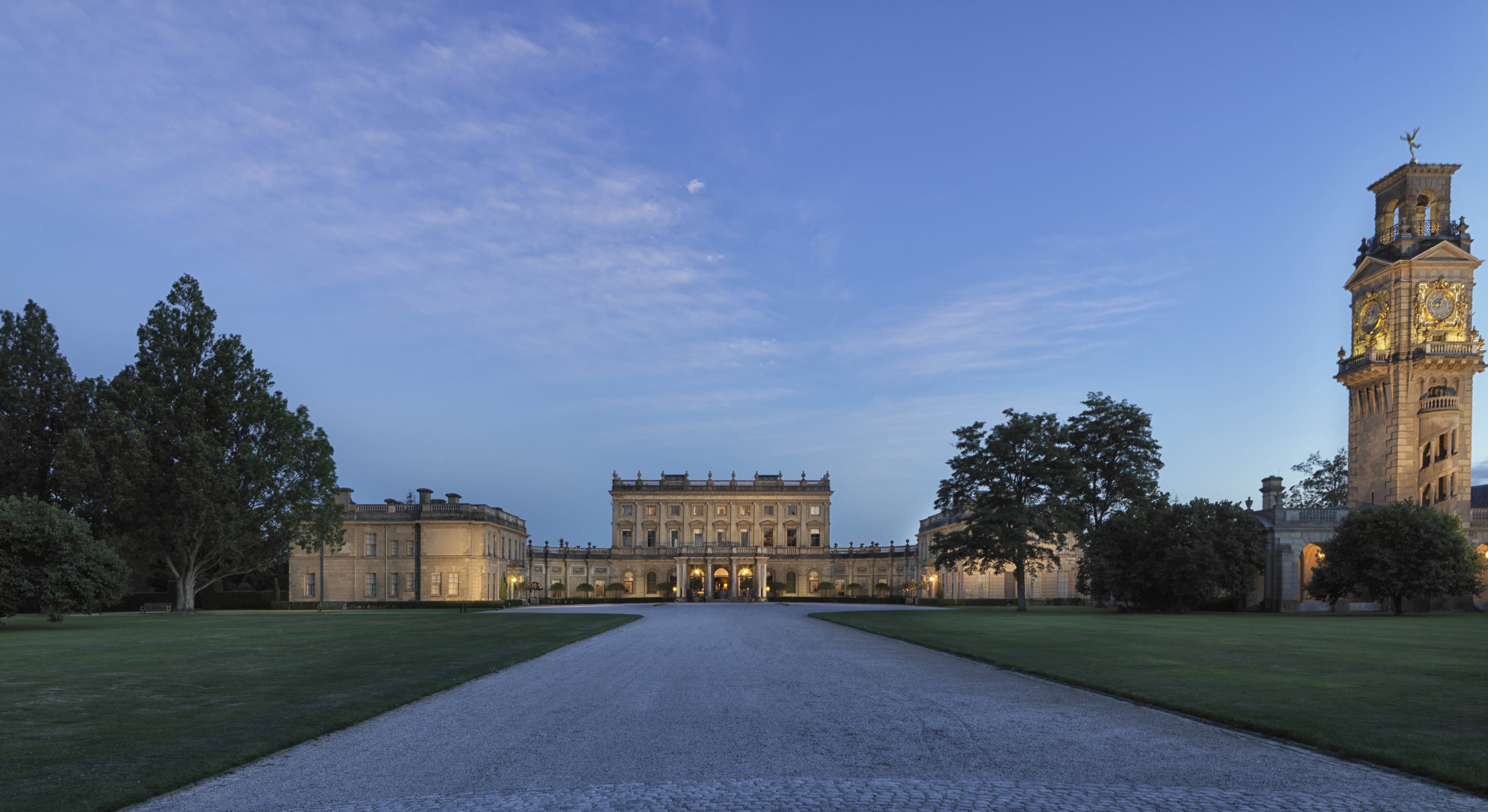 View from driveway of Cliveden House a grand white building with lots of lights on surrounded by grass and trees at night.