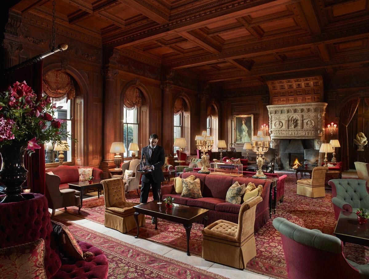 Inside grand luxury room with printed red carpet, velour red sofa's and yellow chairs with large arched windows, a fireplace and huge plant decor.
