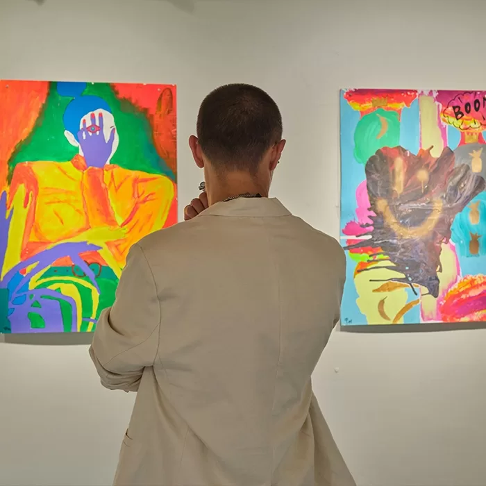 A person with short hair and a beige jacket stands in front of two colorful abstract paintings in an art gallery, viewing the artworks thoughtfully.