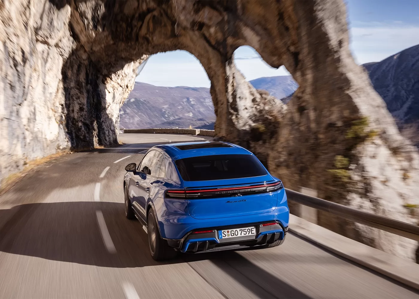A blue SUV drives on a winding mountain road, passing through a natural rock arch with scenic mountains and cliffs in the background.