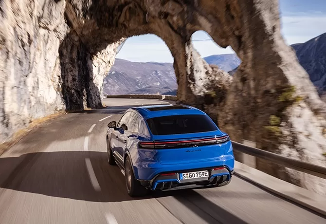 A blue car drives on a winding mountain road, passing through a natural rock arch with scenic cliffs and distant mountains in the background.