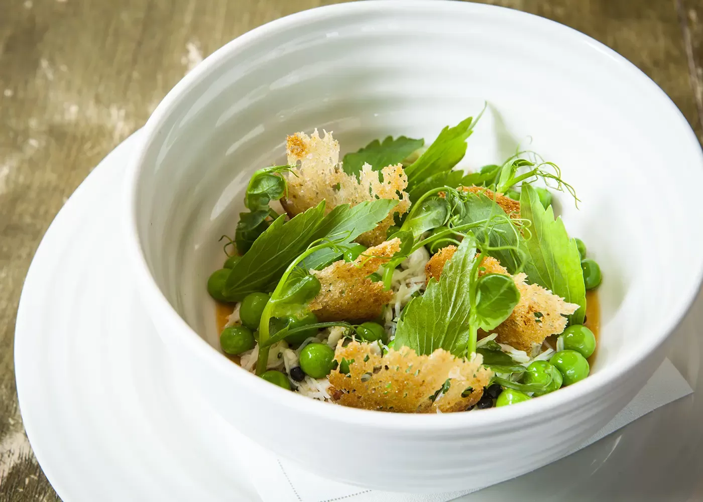 A white bowl filled with fresh green peas, leafy greens, and crispy toasted bread pieces, arranged artfully on a white plate atop a rustic wooden table.
