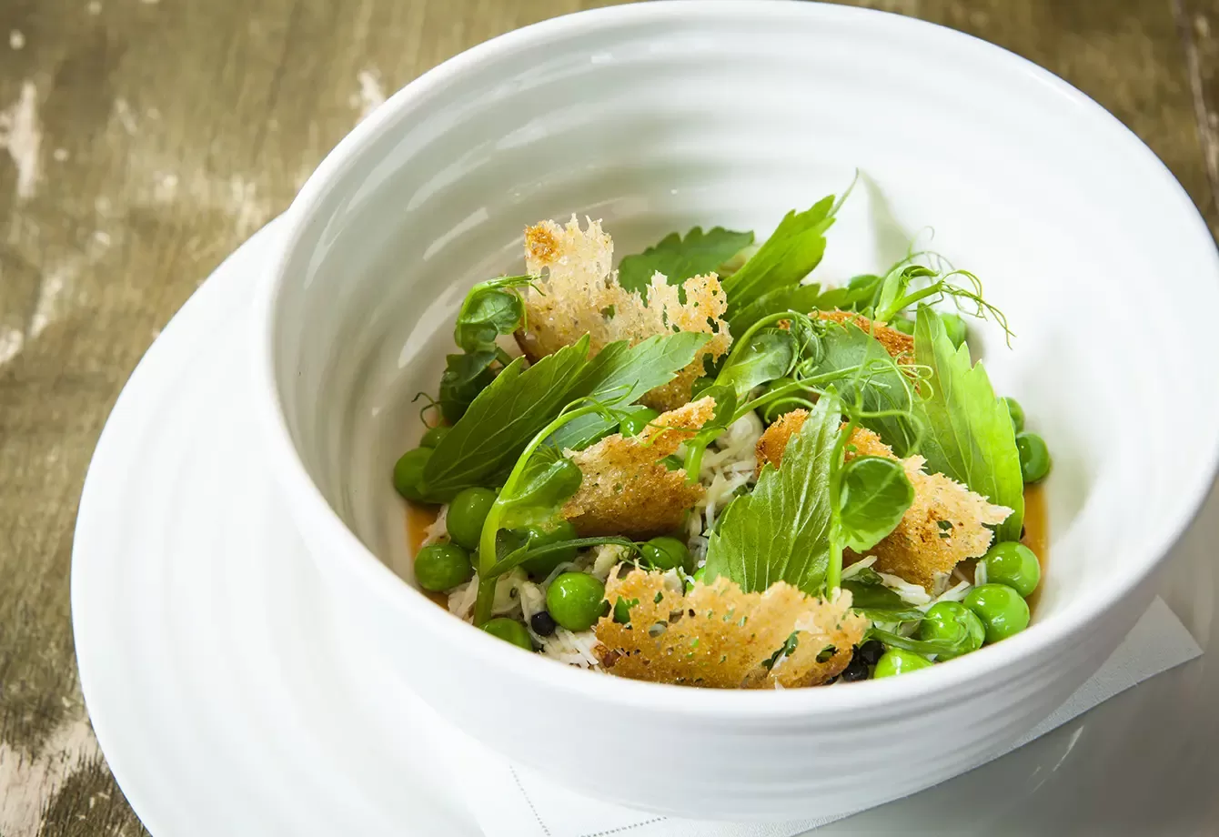 A white bowl contains a salad with green peas, leafy greens, shredded cheese, and crisp pieces of toasted bread, set on a white plate atop a wooden surface.