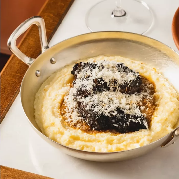 A brass pan filled with creamy polenta topped with braised beef and grated cheese, set on a white surface with part of a wine glass visible in the background.