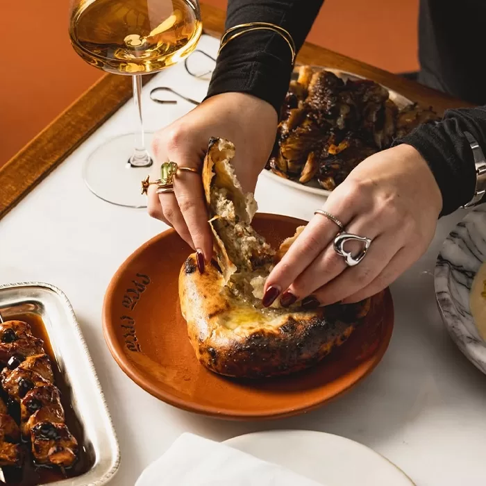 A person tears open a round, baked bread filled with cheese on a brown plate, surrounded by a glass of white wine and other dishes on a white table. The person wears rings and has dark red nail polish.