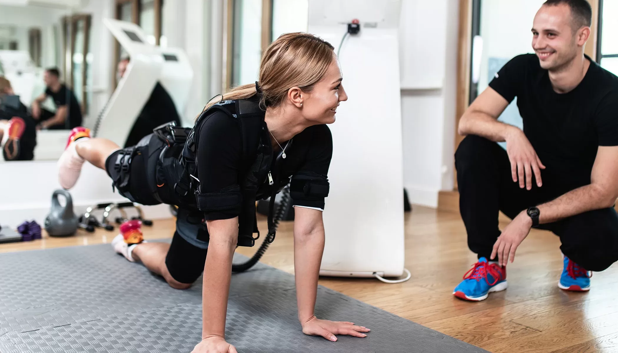 Women on training mats in gym on the floor using a EMS (electro-muscle stimulation) smiling at Personal Trainer