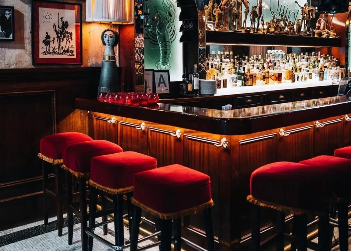 A cosy bar in NYC with a polished wooden counter and red-cushioned barstools. The shelves behind are stocked with various bottles, and decorative art pieces adorn the walls. The ambiance is warm and inviting.