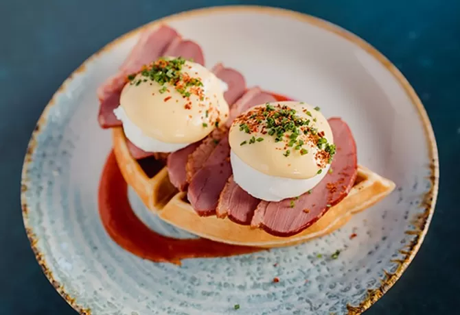 A plate of Eggs Benedict with slices of meat, hollandaise sauce, and herbs on top. The dish is served on a ceramic plate with a decorative rim, sitting on a dark surface at Duck and Waffle, London.