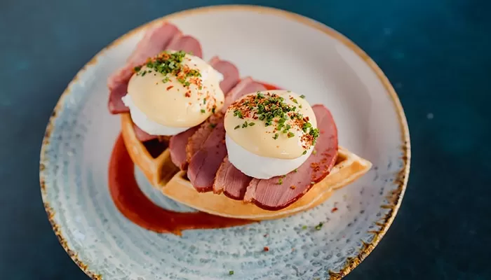 A plate of Eggs Benedict with slices of meat, hollandaise sauce, and herbs on top. The dish is served on a ceramic plate with a decorative rim, sitting on a dark surface at Duck and Waffle, London.