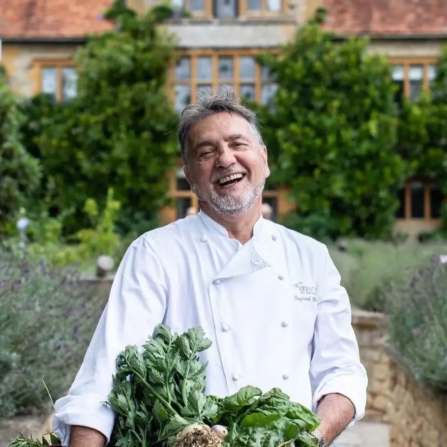 Raymond Blank in his chefs uniform outside of a house carrying a box of vegeatables in a garden