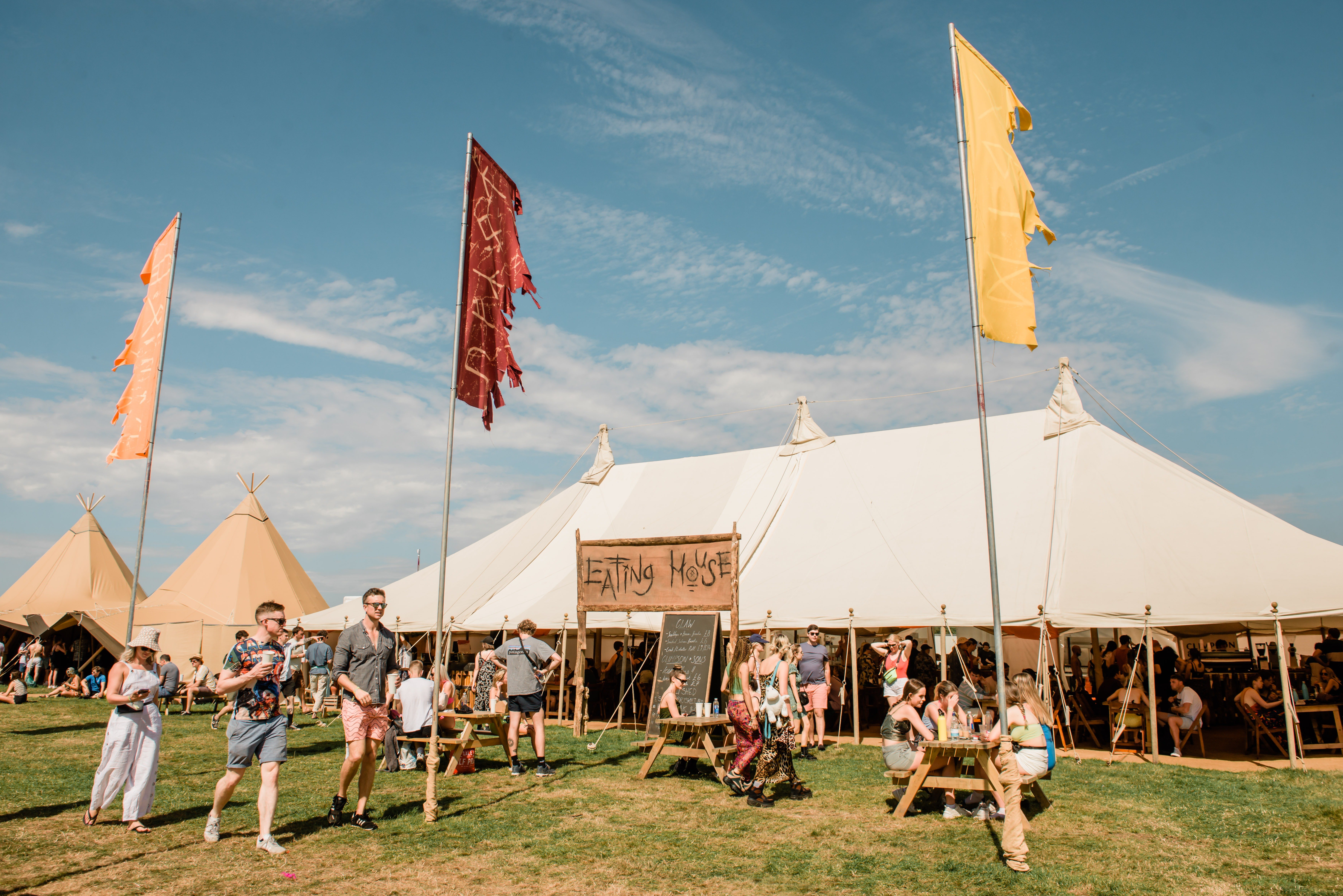 Large white marquee with Eating House sign at a festival with people dining inside, walking outside next to tall bell tents and tall flags.