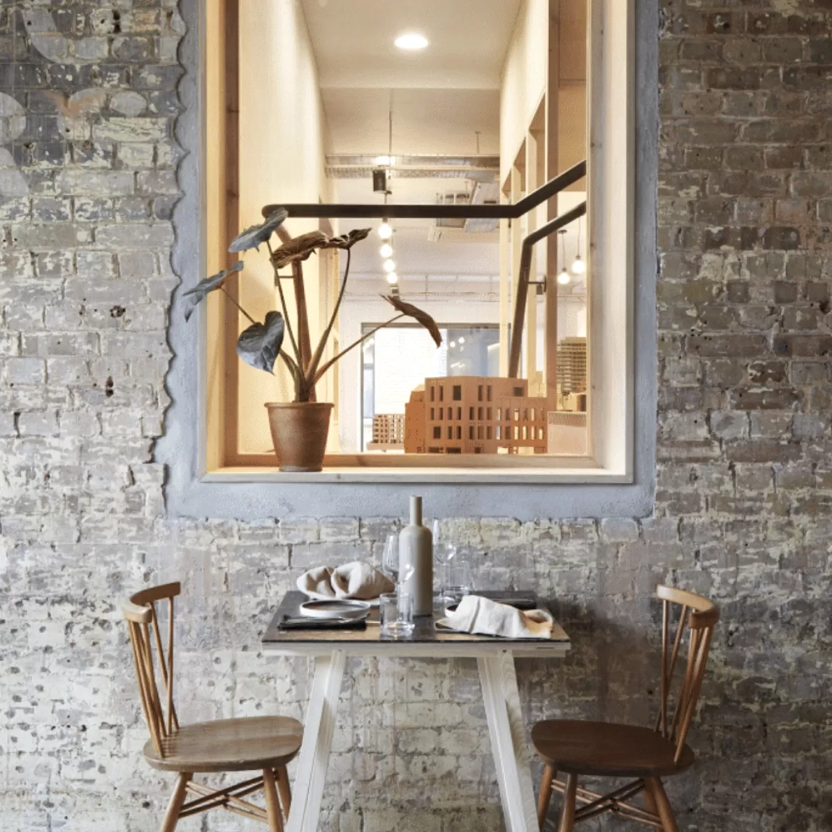 Wooden table of two set up in front of white brick wall with window to upstairs restaurant