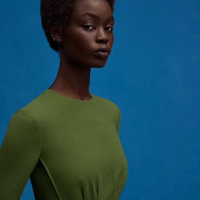 A woman with short curly hair wears a fitted green dress and poses against a solid blue background, looking confidently towards the camera.