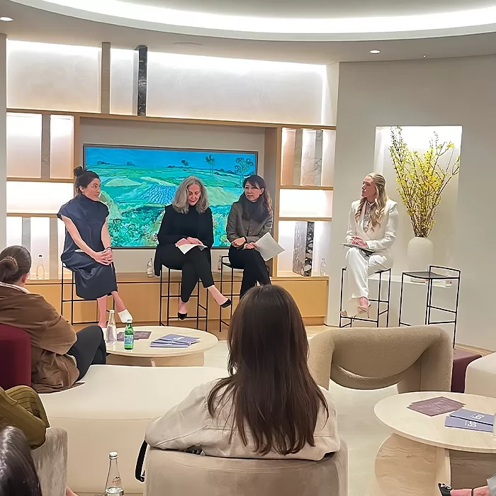 Four women are seated at the front of a modern, well-lit room, engaging in a panel discussion while an audience listens. A colorful landscape painting and flower arrangement are in the background.