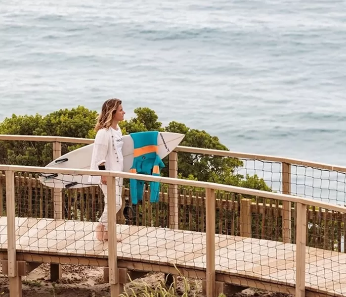 Women stood on a wooden walkway bridge across beach next to the sea holding a surf board at Ericeira, Portugal