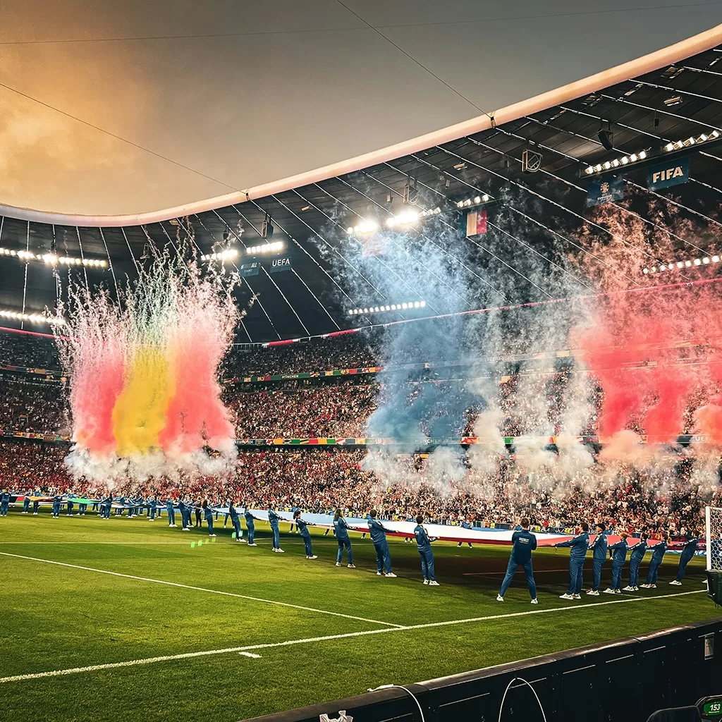 A stadium filled with cheering fans during a soccer match, with colorful smoke and confetti—red, yellow, and blue—shooting into the air as uniformed staff stand along the field holding a large flag.