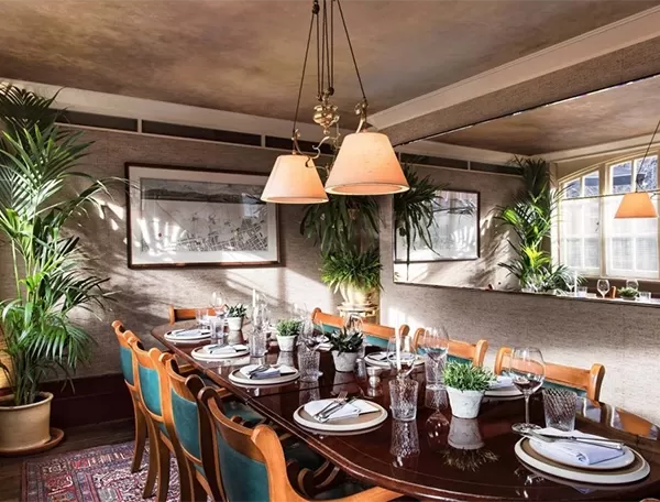 A warmly lit dining room with a long wooden table set for eight. Green upholstered chairs surround the table, which is adorned with plants and dinnerware. Large windows and wall art add to the cosy atmosphere at Chiltern Firehouse, London.