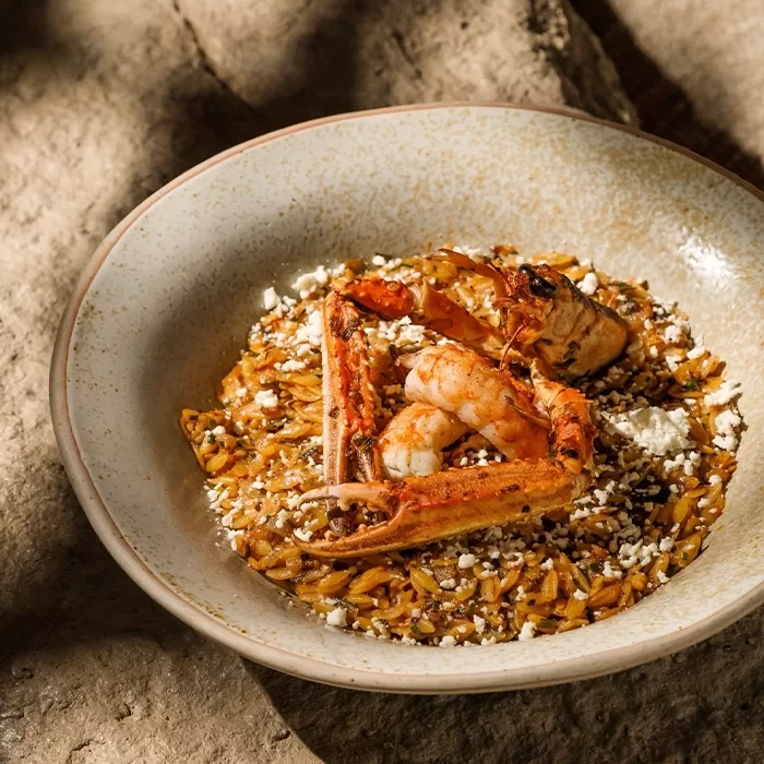 A bowl of orzo pasta topped with grilled shrimp and shellfish, garnished with crumbled white cheese, served on a rustic ceramic plate placed on a textured stone surface.