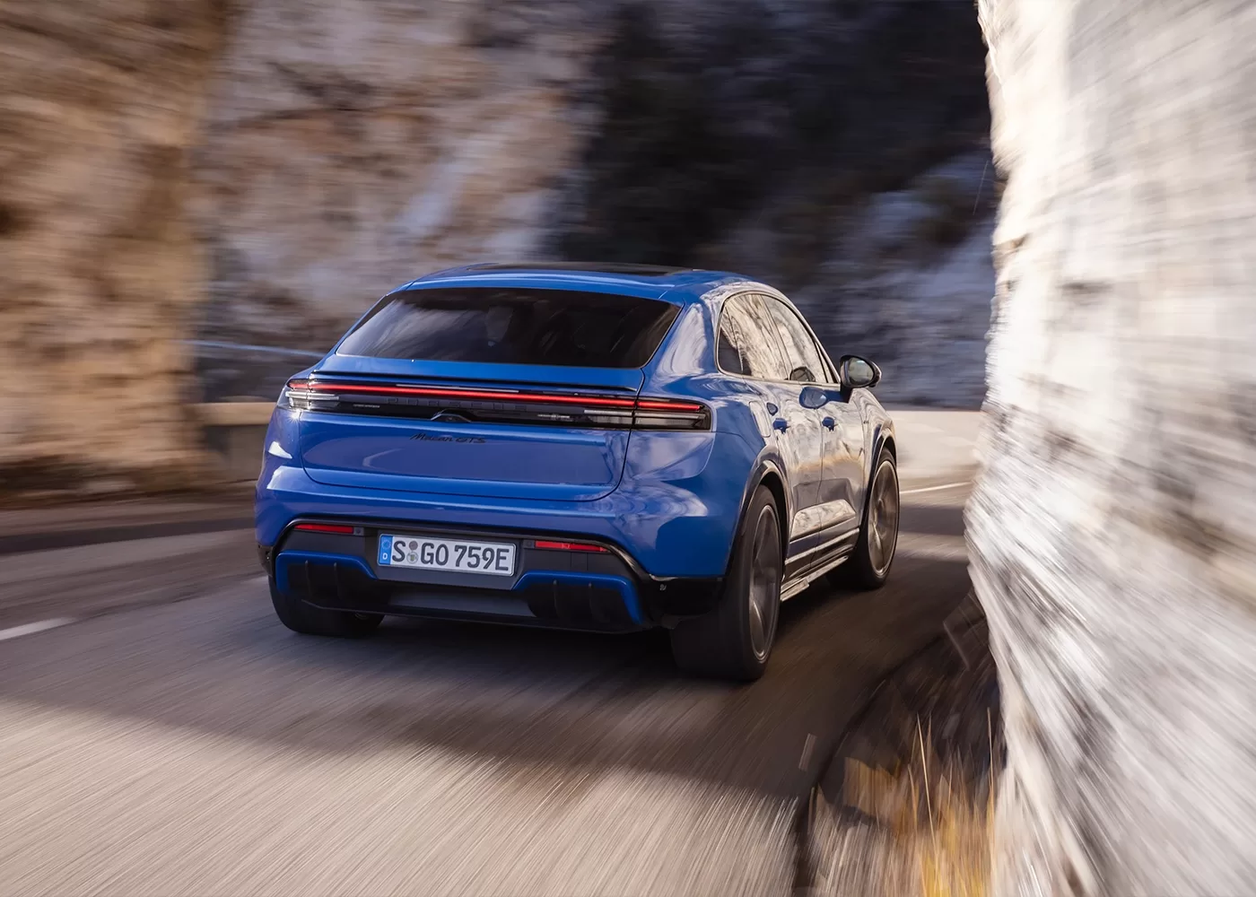 A blue electric SUV is driving on a winding mountain road, passing closely by a rocky cliffside. The scene is in motion, and the vehicles rear and taillights are visible.