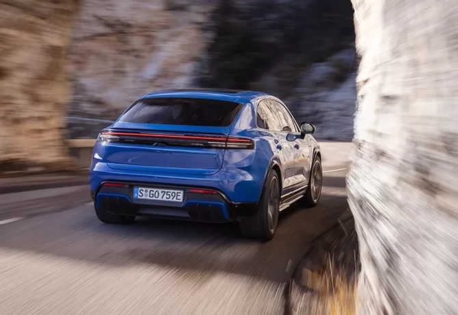 A blue sports SUV drives along a winding mountain road, passing close to rocky cliffs. The car is in motion and the road appears curvy, suggesting speed and dynamic movement.