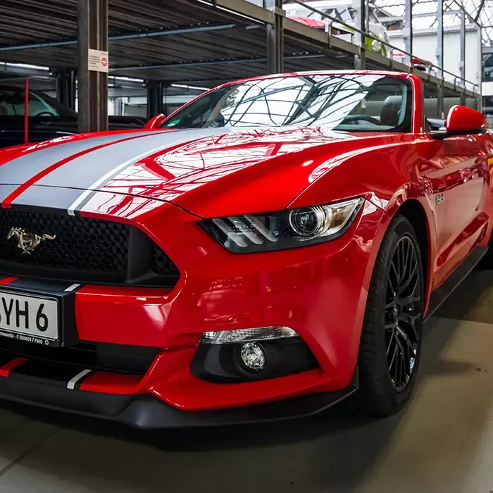 A bright red Ford Mustang sports car with black racing stripes is parked indoors under a metal structure, reflecting overhead lights on its hood and windshield. The license plate reads “SYH 6.”.