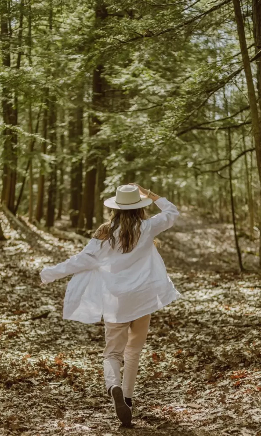 A person wearing a white shirt, light pants, and a hat walks along a wooded trail, surrounded by trees and dappled sunlight filtering through the leaves.