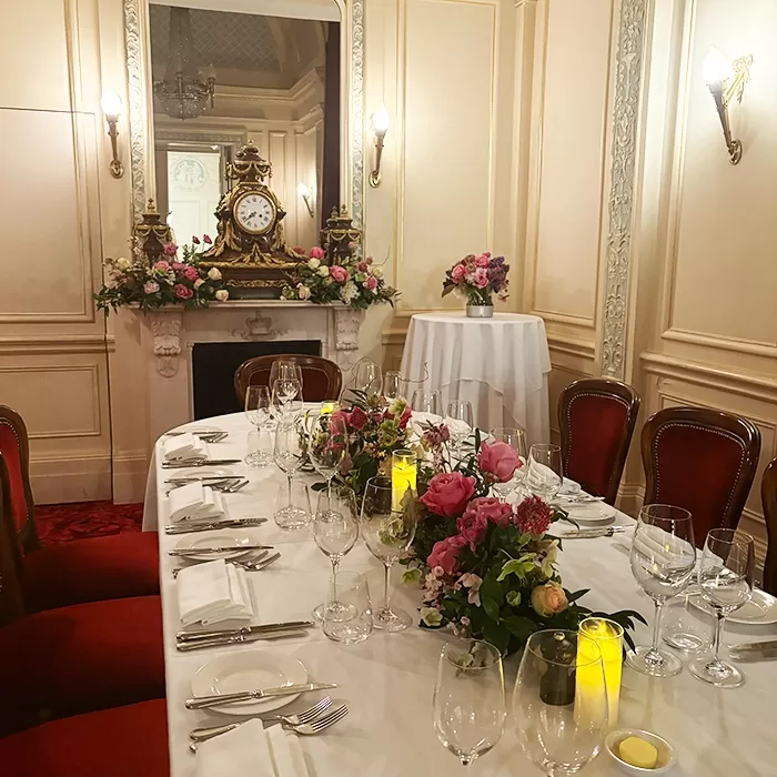 Elegant dining room with a long table set for a formal meal, featuring red chairs, floral centerpieces, candles, and a large ornate clock on a mantel with more flowers in the background.