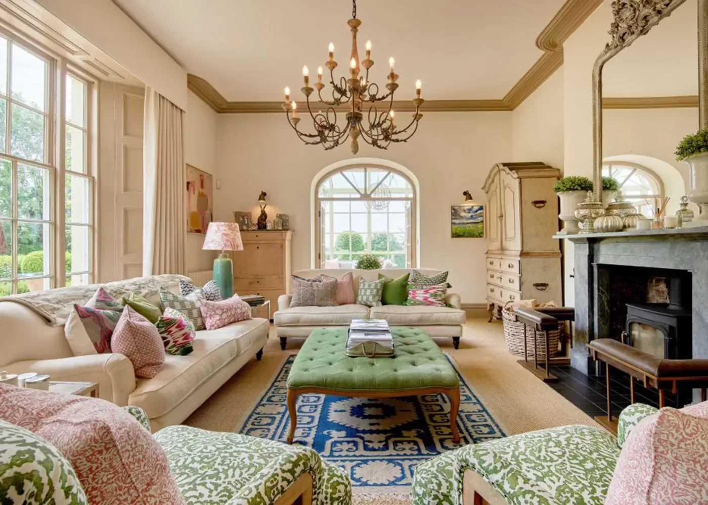 Elegant living room with large windows, plush sofas and armchairs with patterned cushions, a green tufted ottoman, blue rug, fireplace, chandelier, and classic decor accents. Bright and cozy atmosphere.