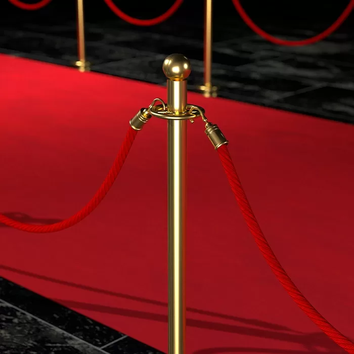 A close-up of a gold stanchion with a red velvet rope on a red carpet, with a dark tiled floor and more stanchions and ropes in the background.