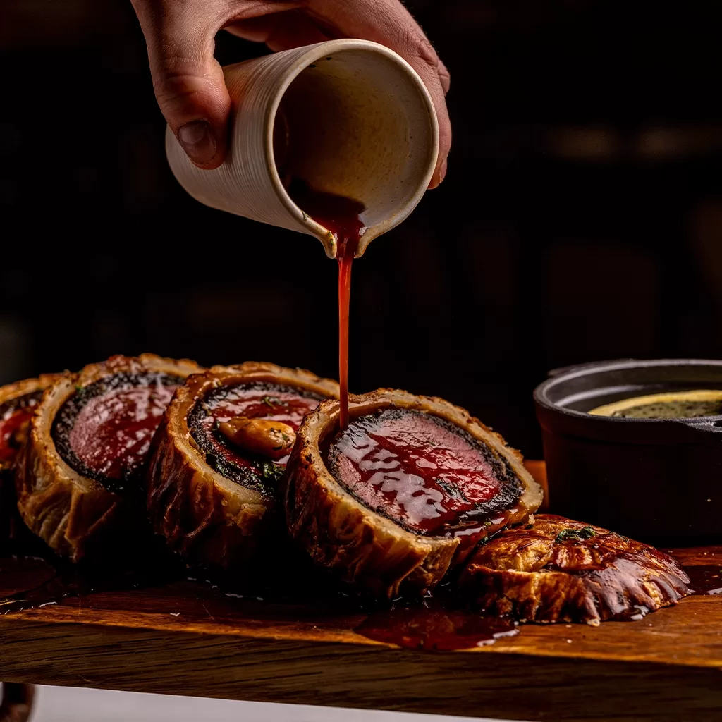 A hand pours sauce over sliced Beef Wellington on a wooden board, with a small pot of extra sauce nearby. The dish reveals tender beef wrapped in pastry and mushroom duxelles.