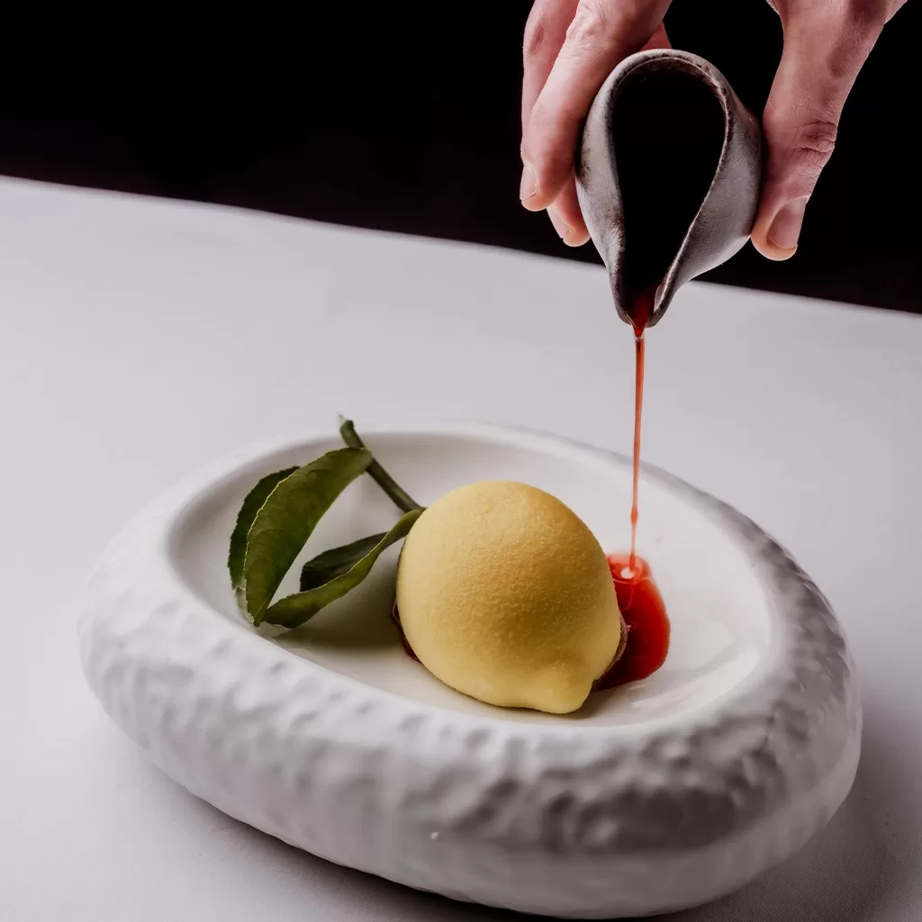 A hand pours red sauce from a small pitcher onto a dessert shaped like a lemon, garnished with green leaves, served in a textured white dish on a white tablecloth.