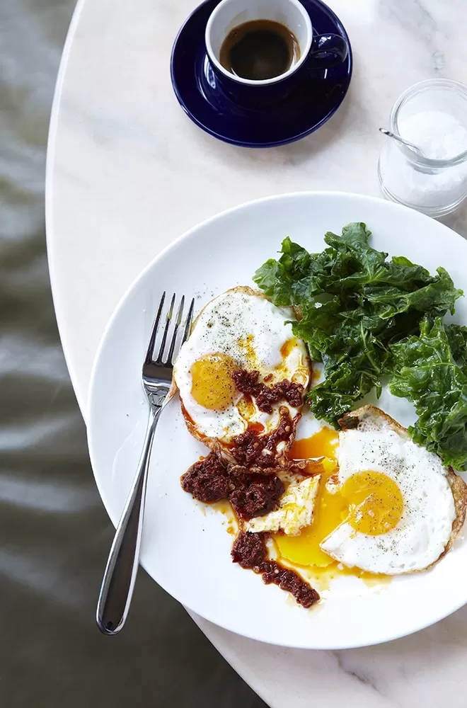 A breakfast plate with two sunny-side-up eggs, greens, and a mound of sauce on a white plate, next to a fork. A small cup of coffee and a glass of cream are on the table, which has a marble-like surface
at Granger & Co, London.