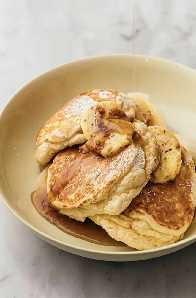 A plate of fluffy pancakes topped with sliced bananas and drizzled with syrup sits on a light-colored surface. The syrup glistens on the pancakes, enhancing their golden-brown texture at Granger & Co, London.