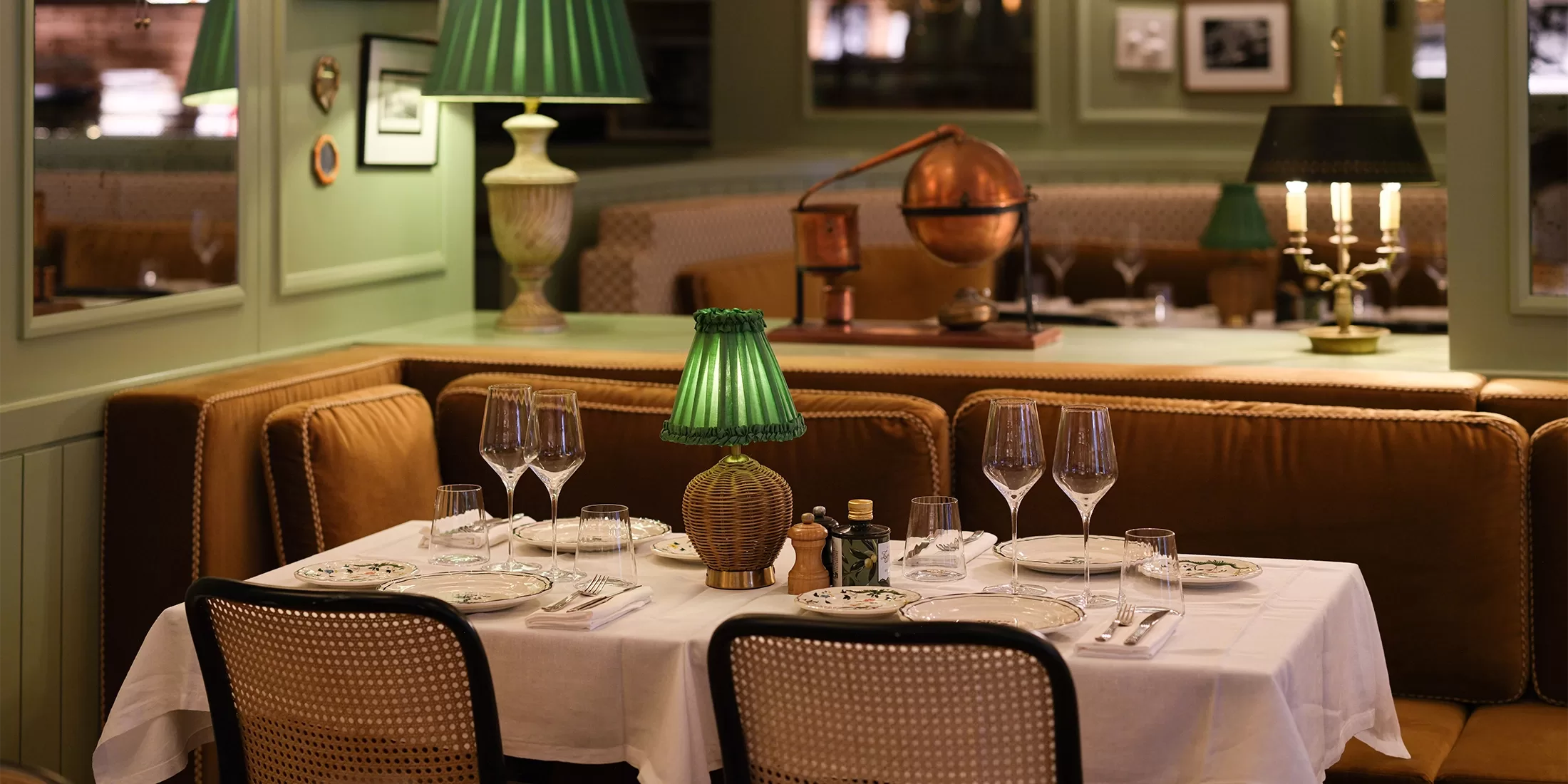 An indoor restaurant scene with an intimate and cozy ambiance. The walls are painted in soft green, and the space is warmly lit with soft lighting. The dining area features plush brown seating, and the tables are neatly set with white tablecloths, wine glasses, and cutlery. A vintage green lamp stands on the table, adding a classic touch. The decor includes framed black-and-white photographs on the walls, along with a mix of modern and antique furnishings at Zeffirino, Monte Carlo.