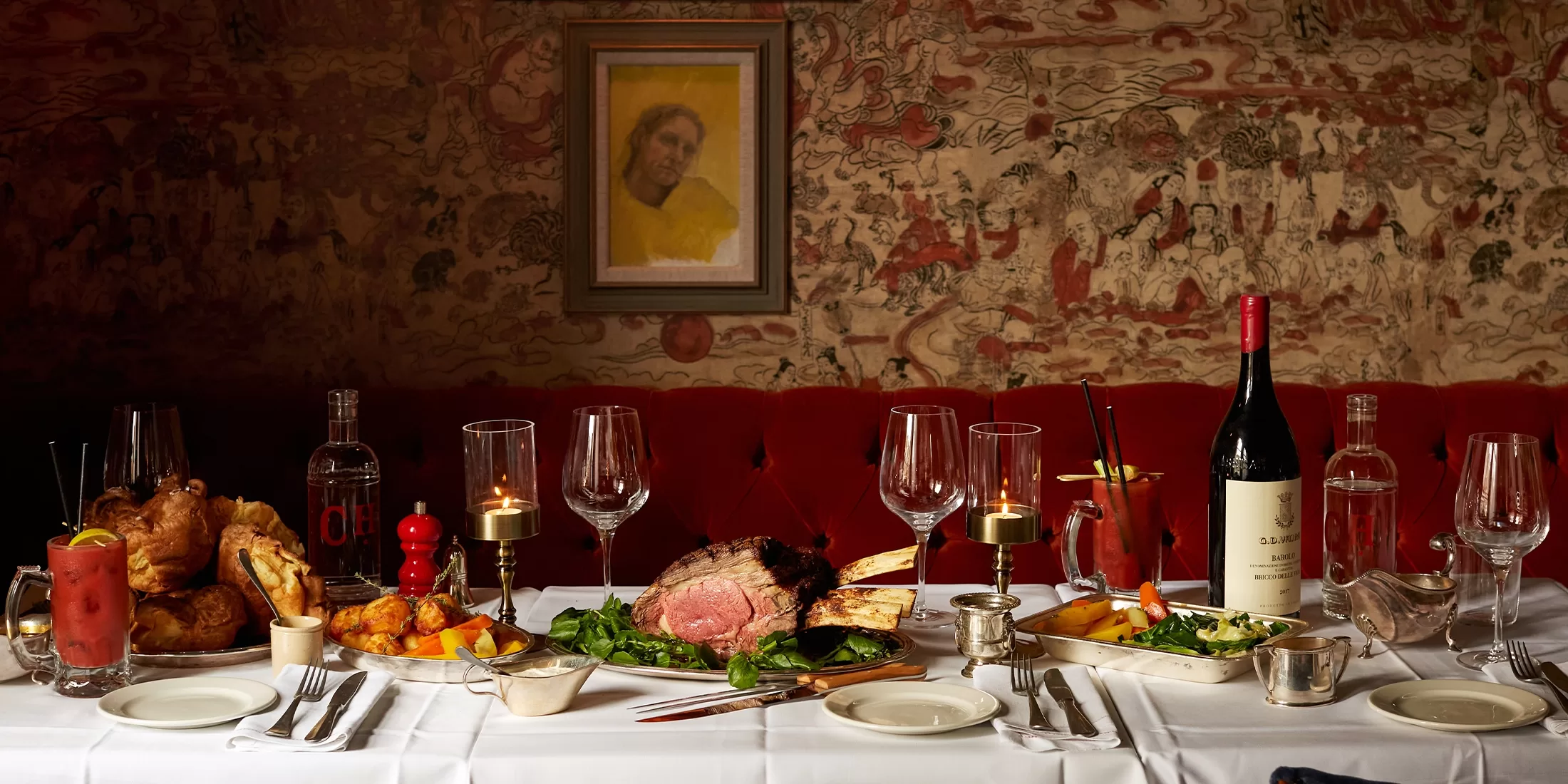 A white tablecloth dinner table is set with roast beef, Yorkshire pudding, roast potatoes, vegetables, wine, cocktails, and candles. A red velvet booth and tapestry wall with a portrait are in the background.
