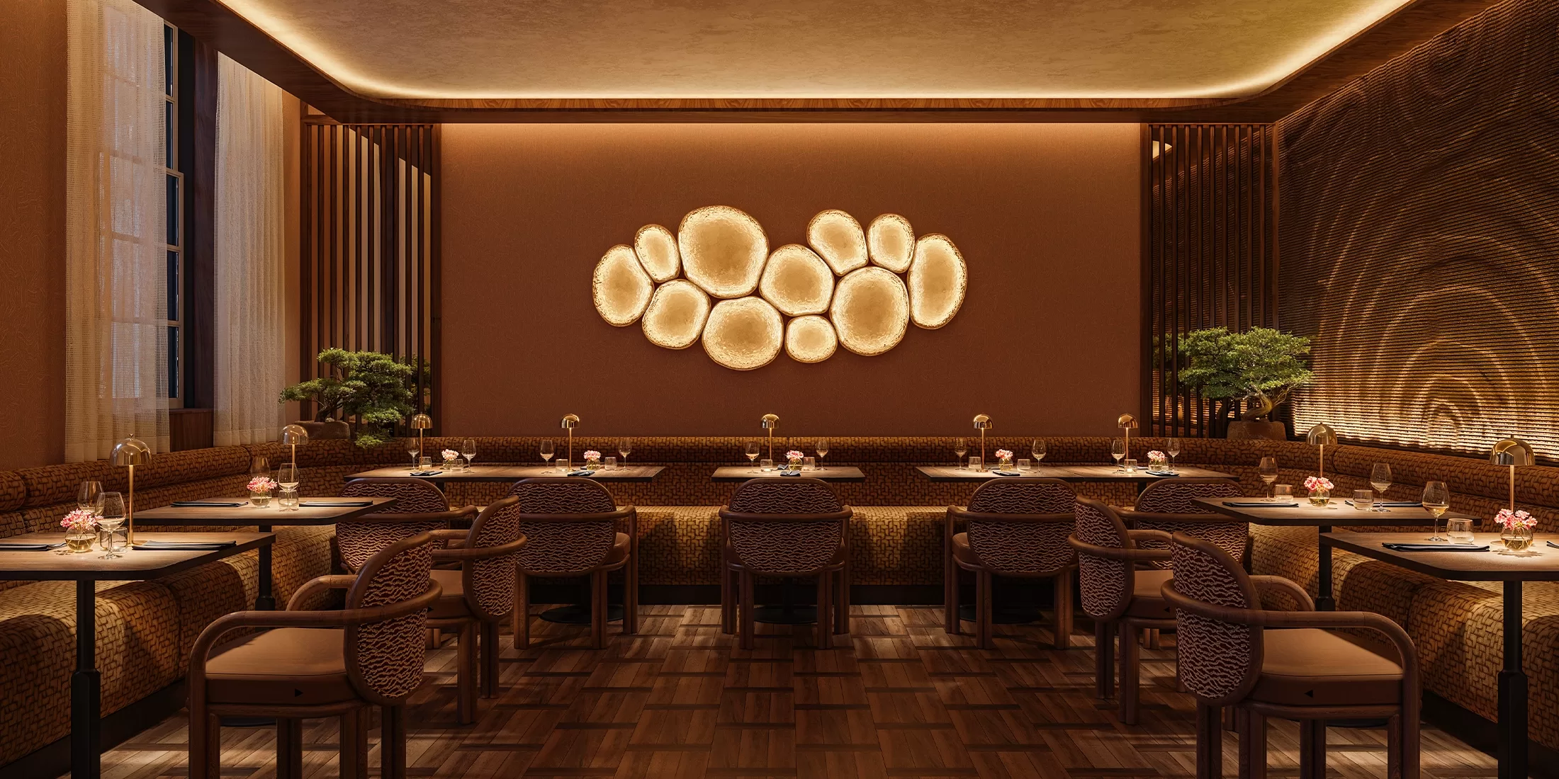 A modern, warmly lit restaurant with brown walls, woven chairs, wooden tables, and a glowing circular wall art centerpiece. Small plants and floral arrangements decorate the tables and corners.