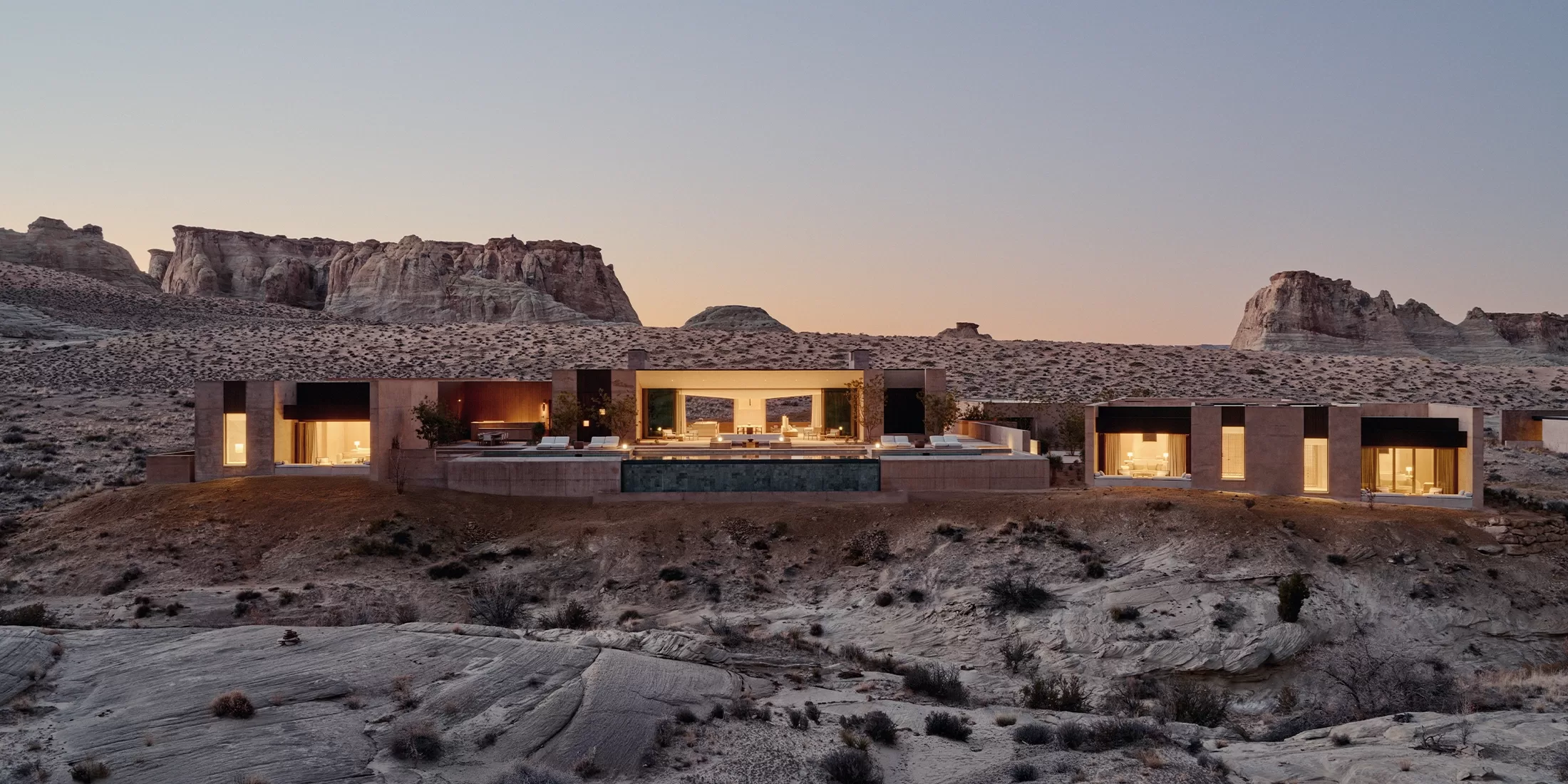 A modern, luxurious house with large glass windows is set in a rocky desert landscape at dusk. The interior lights glow warmly, and a central swimming pool faces the arid terrain and distant cliffs.
