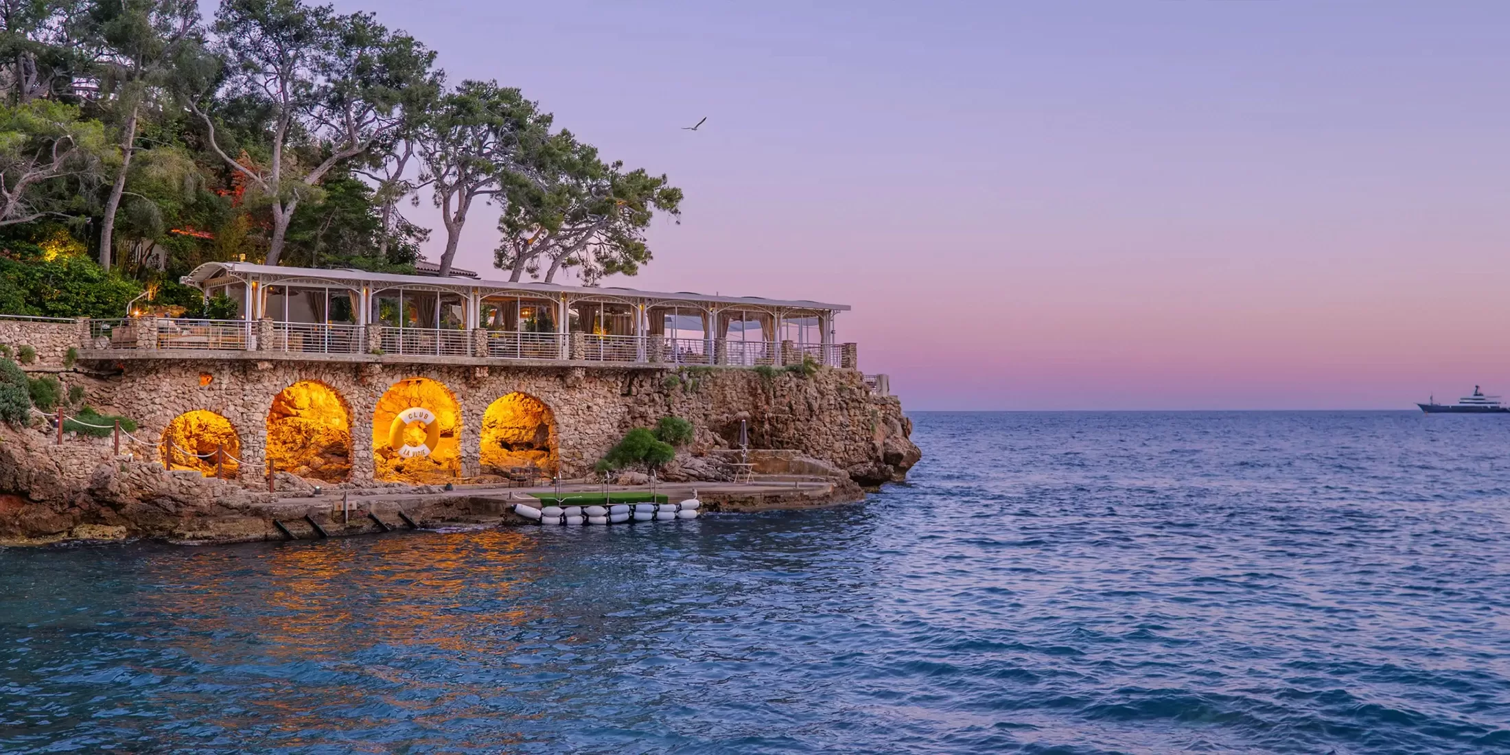 A stone building with arched entrances and warm lights sits atop a rocky shoreline at sunset, surrounded by trees. Calm blue sea and a yacht are visible, with a pastel pink and purple sky in the background.