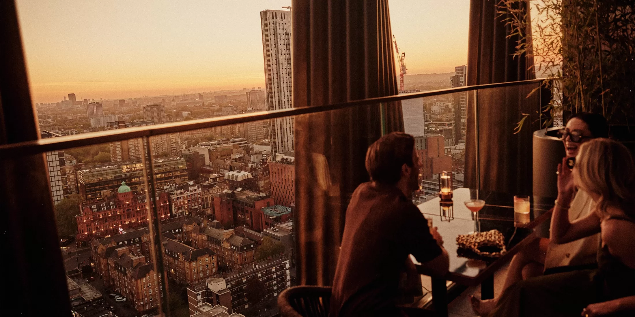 Three people sit at a rooftop bar table at sunset, enjoying drinks and snacks, with a city skyline and tall buildings visible in the background through glass panels.