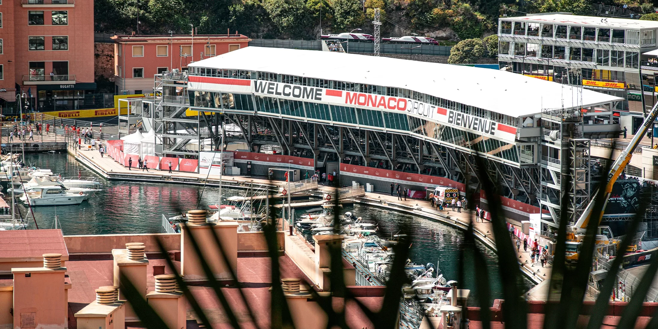 A large Welcome to Monaco sign is displayed above a grandstand overlooking a marina with yachts and boats, surrounded by buildings and greenery. The scene is partially framed by out-of-focus plants in the foreground.