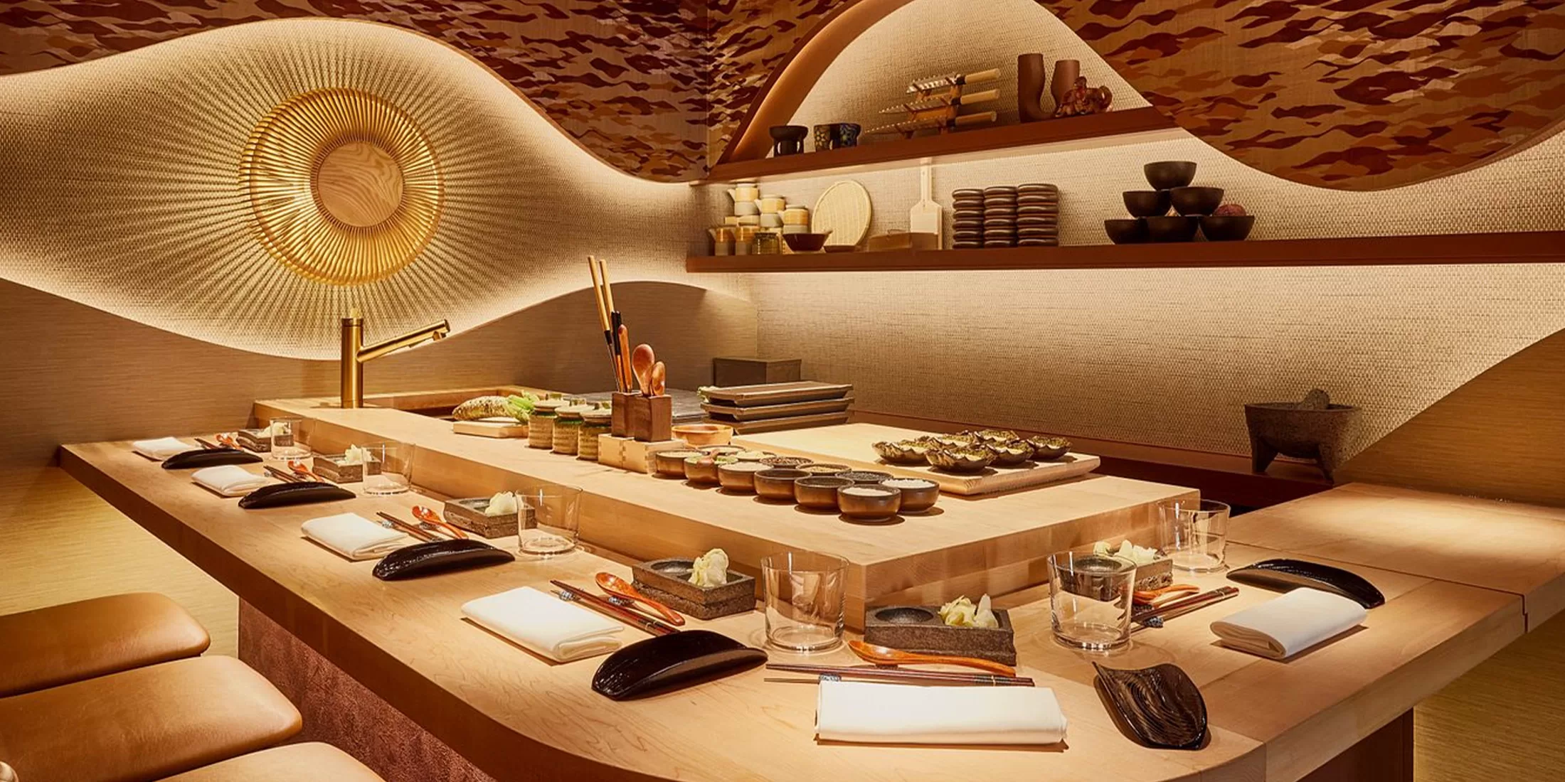 A minimalist yet luxurious sushi bar with a sleek, modern design at Juno Omakase, London. The dining area is set with wooden tables, seating for guests, and a stunning curved, illuminated wall accentuating the space. Plates, chopsticks, and sushi rolls are meticulously arranged on the bar.