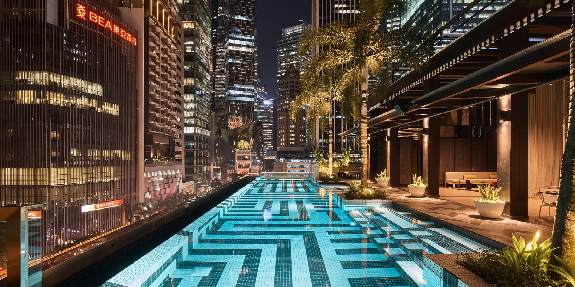 A rooftop pool at night with geometric tile patterns, surrounded by modern buildings. The area is lit with warm lights, and there are seating areas with plants. Skyscrapers loom in the background, adding an urban atmosphere.