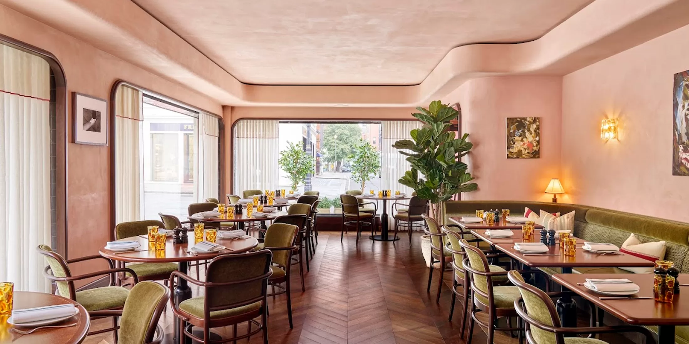 A chic restaurant interior with pink walls and a large window. Tables are set with yellow glassware and napkins, surrounded by green velvet chairs. Plants and soft lighting add a cozy, elegant ambiance.