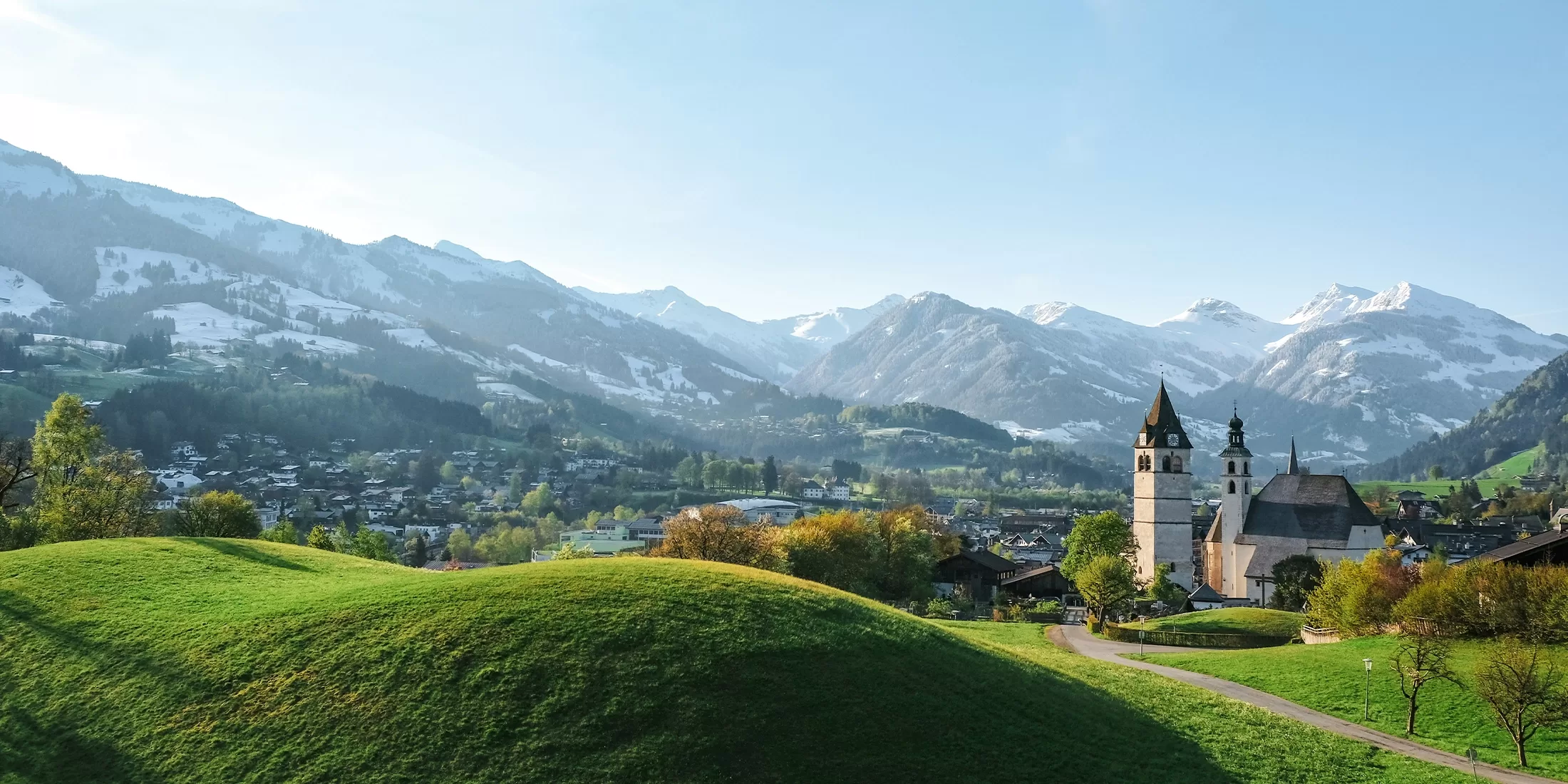 A picturesque village in Kitzbühel nestled in a lush green valley, with a church and scattered houses. Snow-capped mountains frame the background under a clear blue sky, creating a serene and idyllic landscape.